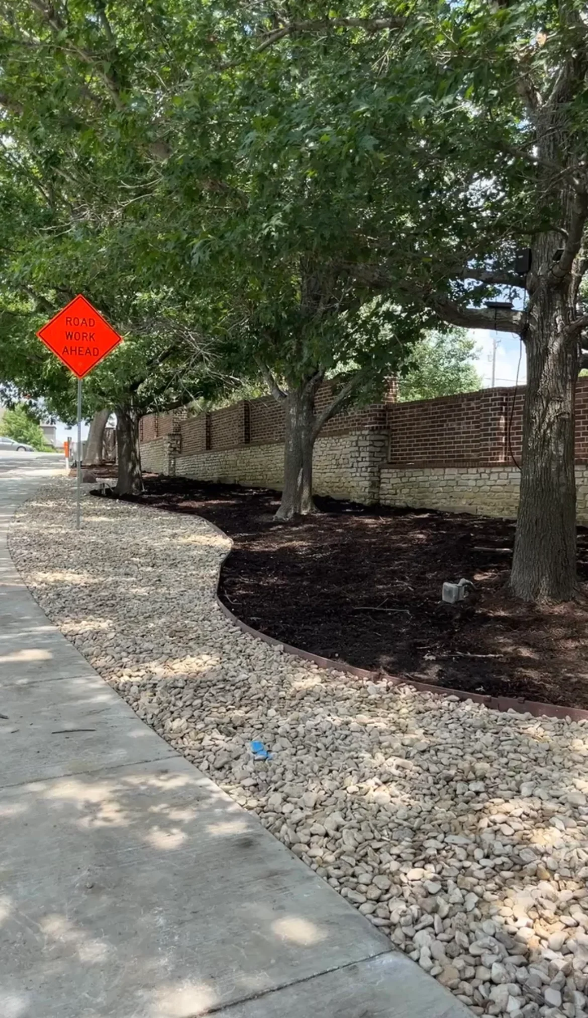 Landscape beds with dark mulch and light stone edging around mature trees against brick wall